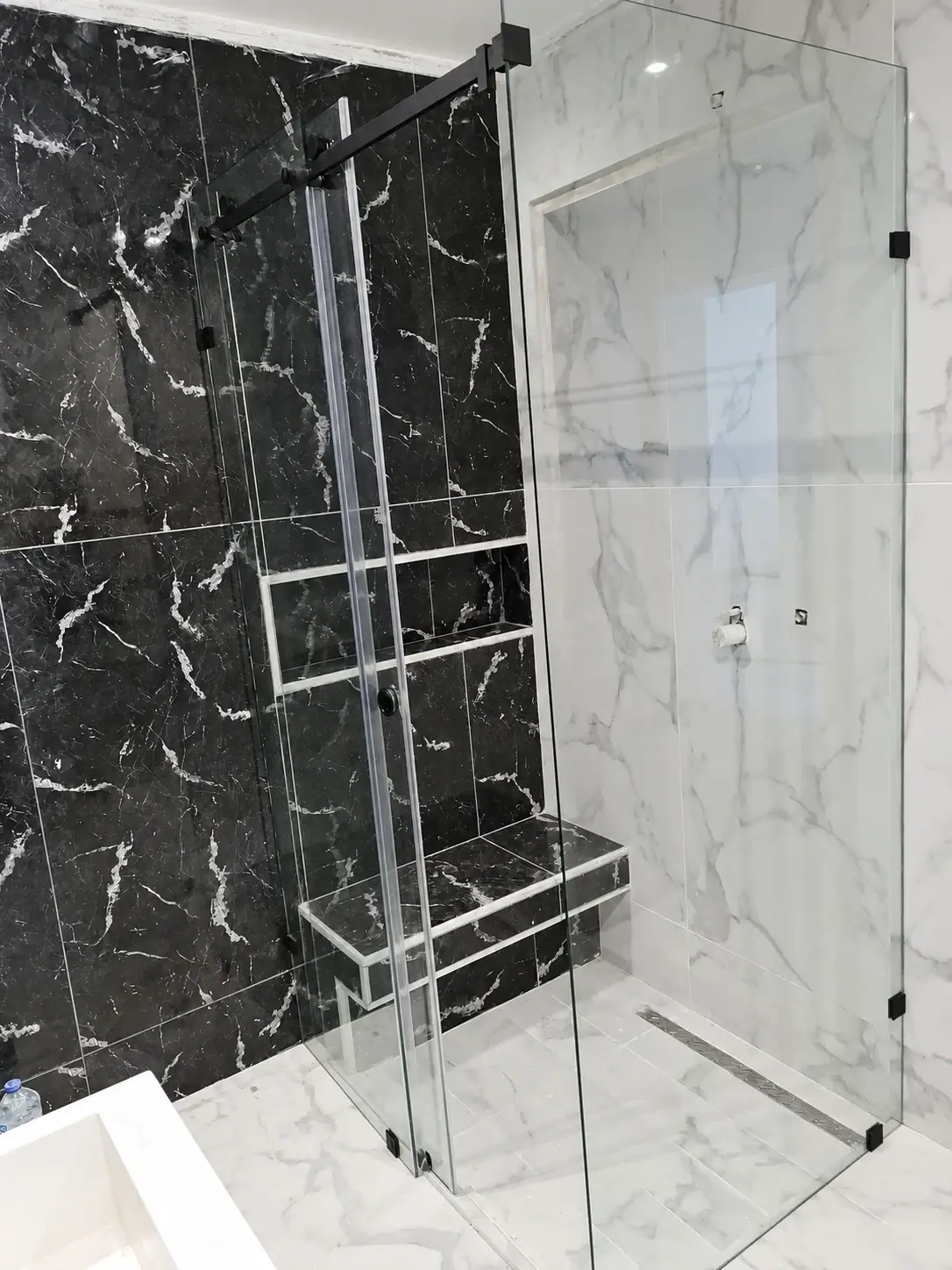 Cabine de douche en verre clair avec porte coulissante sur rail noir, dans une salle de bain en marbre noir et blanc