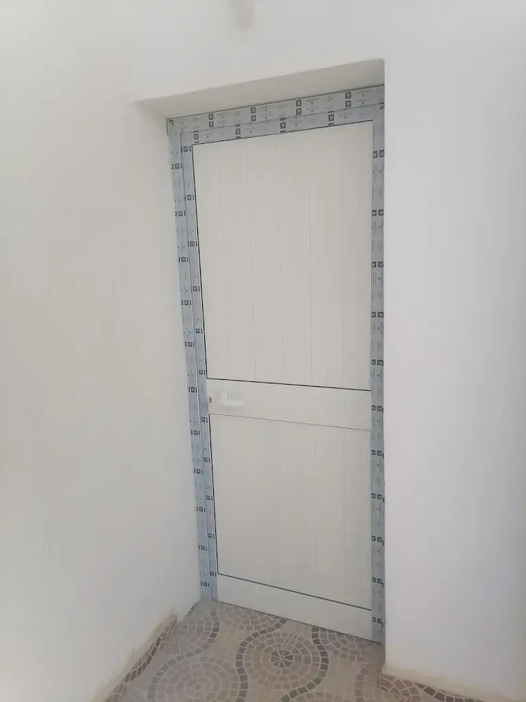 Porte de service en aluminium blanc à lames verticales, encadrement encore protégé par le film de pose