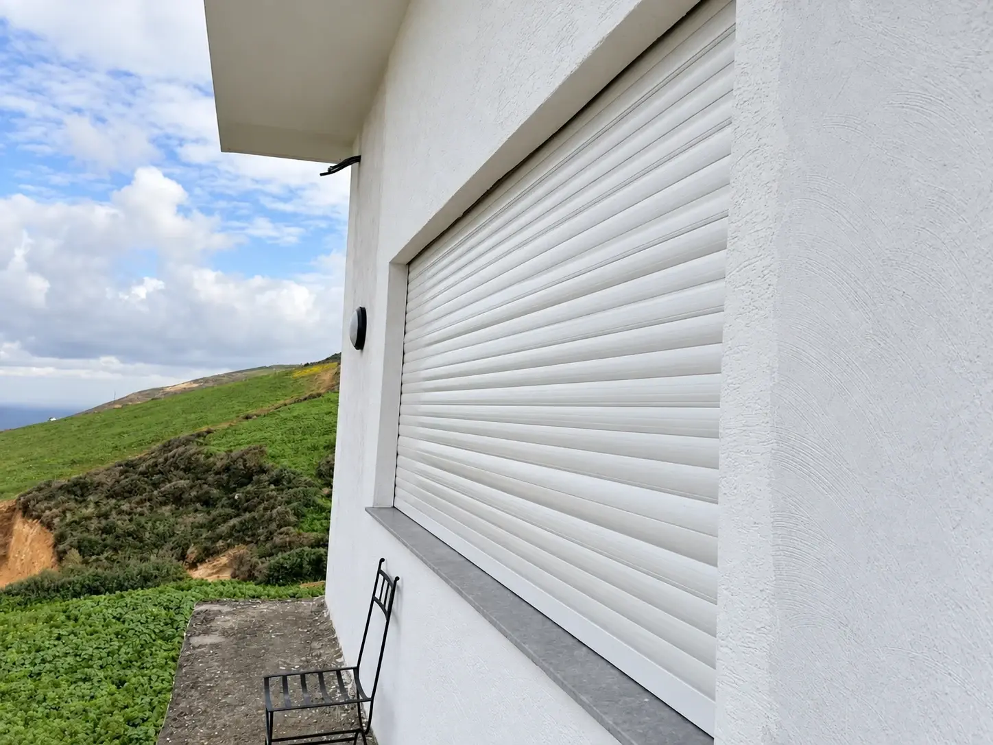 Volet roulant en aluminium blanc fermé sur la fenêtre d'une maison, face à la colline et à la mer