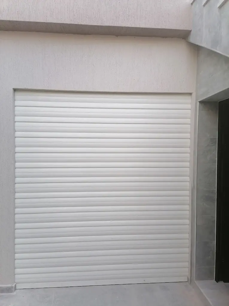 Volet roulant en aluminium blanc fermant une grande ouverture de garage, à côté d'une entrée carrelée