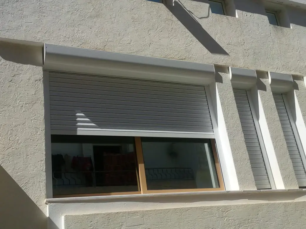 Volet roulant en aluminium à coffre extérieur blanc, à moitié ouvert sur une fenêtre coulissante aspect bois